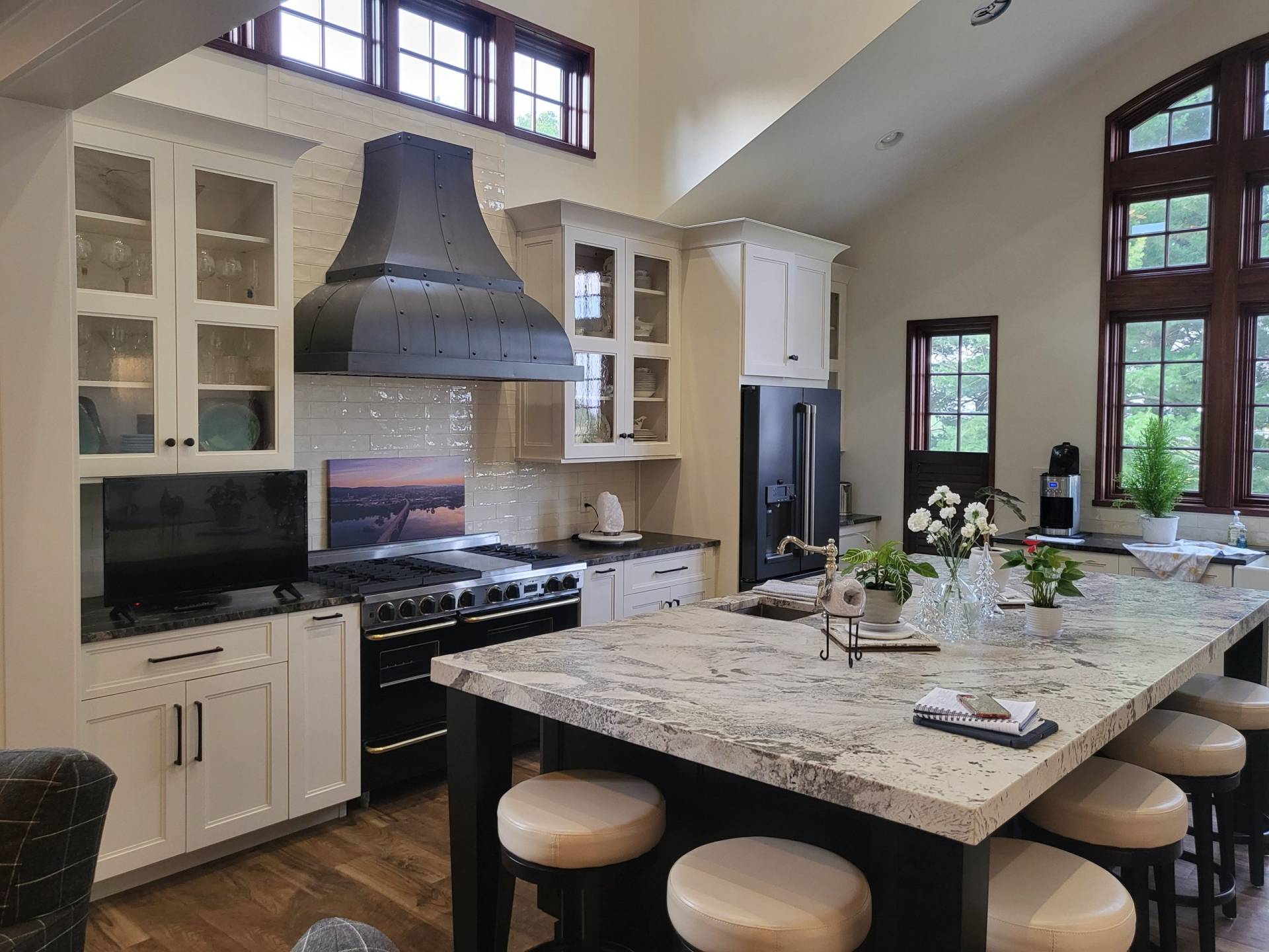 Interior photo of custom kitchen with cathedral ceilings, extra large granite countertop island, and custom hood over luxury range with white cabinets. Windows have natural burgundy wood trim, letting in abundant natural light. Designed by Brad M. Zahm, RA, NCARB and The Mula Group.