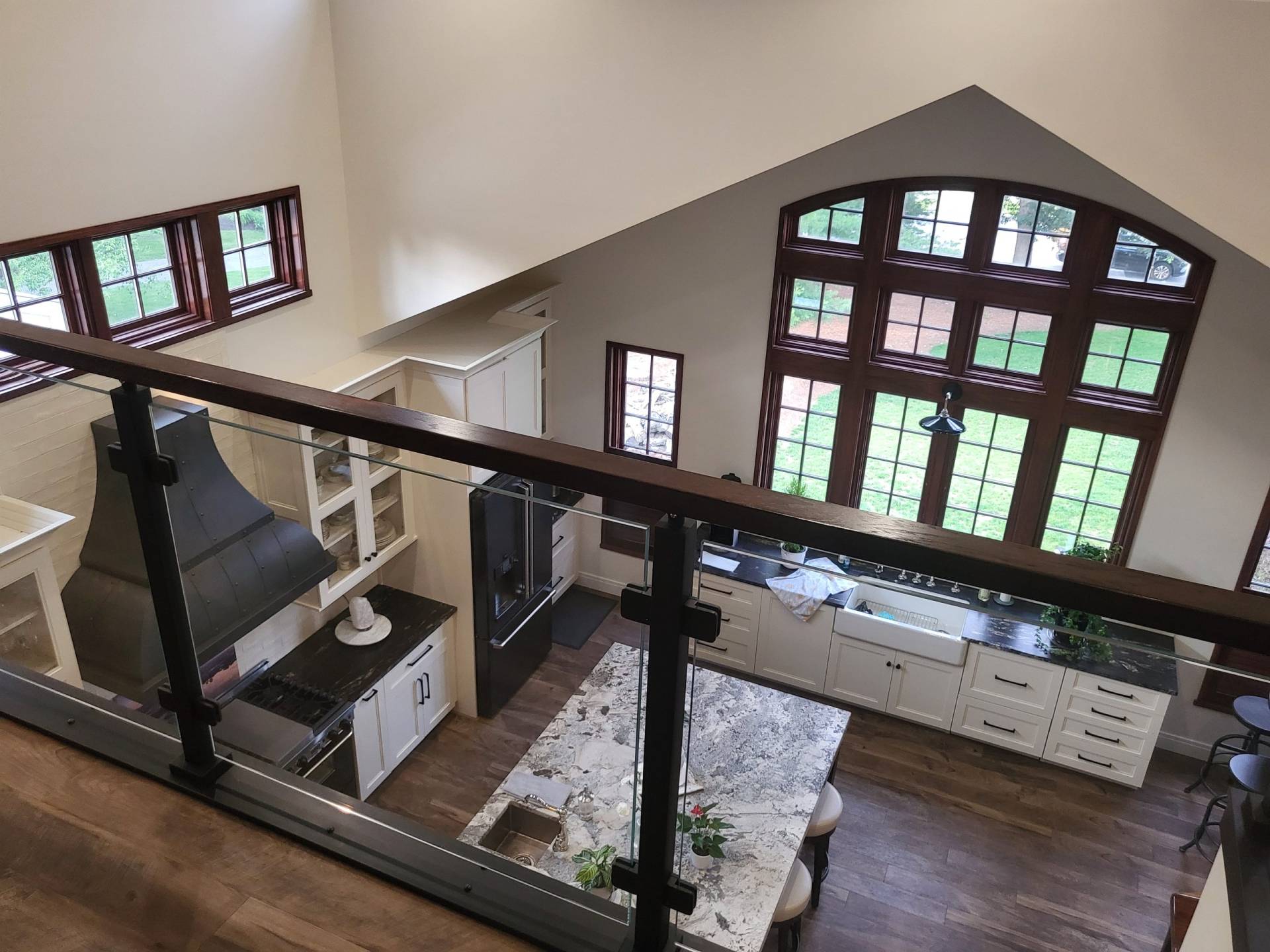 Second floor view from library of custom kitchen with cathedral ceiling and granite countertop over white cabinets. Large arched windows with natural burgundy trim fill the wall. Glass railing allows uninterrupted sightline. Designed by Brad M. Zahm, RA, NCARB and The Mula Group