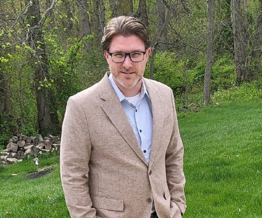 Brad M. Zahm, RA, NCARB wearing gray slacks and shoes with a button-down shirt and tan linen blazer standing on a concrete patio with budding trees and shrubs in the background.
